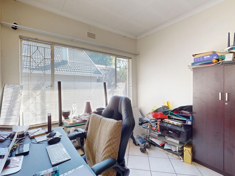 A functional home office space featuring tiled flooring, a large window with venetian blinds, and neutral wall finishes.