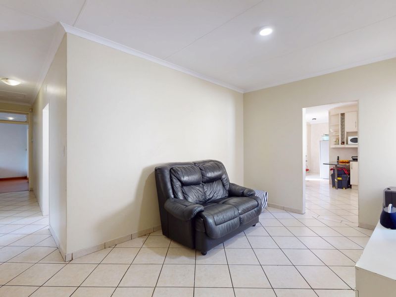A living room area featuring neutral tiled flooring and recessed lighting, with an open-plan layout providing a view into the adjacent kitchen space.