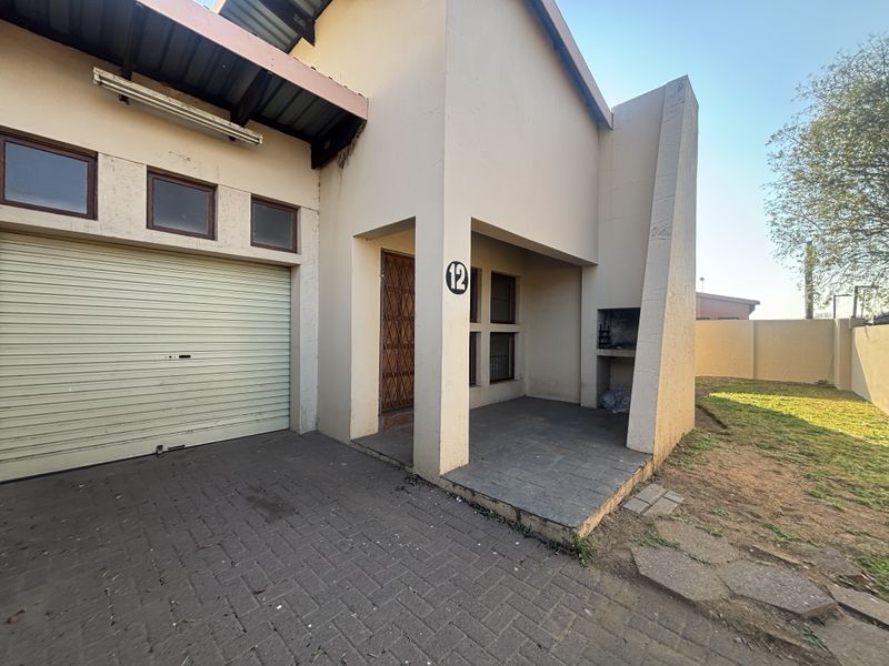 Front exterior view of a townhouse featuring a single garage, paved driveway, and a covered patio area with a built-in braai.