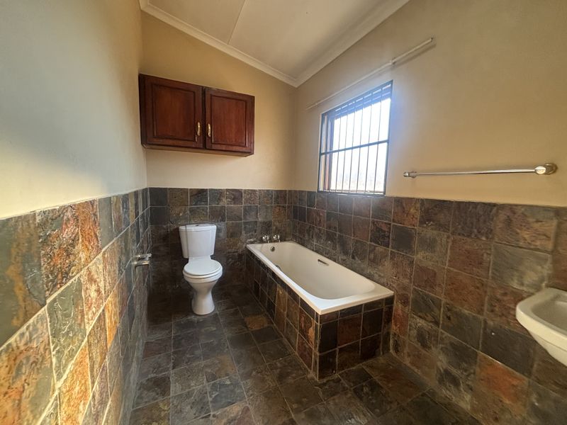 A functional bathroom featuring a built-in bathtub, toilet, and wall-mounted storage cabinet, finished with rustic slate-style wall and floor tiles.