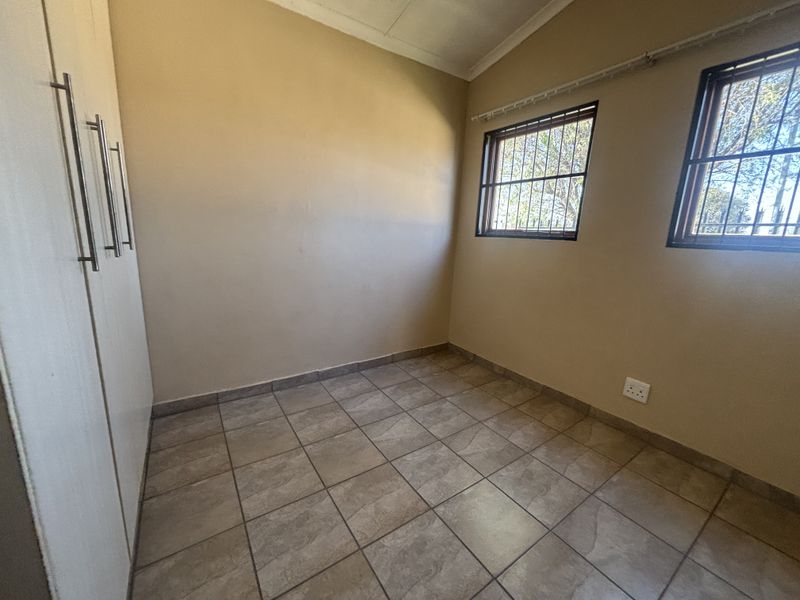 A clean, empty bedroom featuring tiled flooring, built-in cupboards, and windows fitted with security bars.