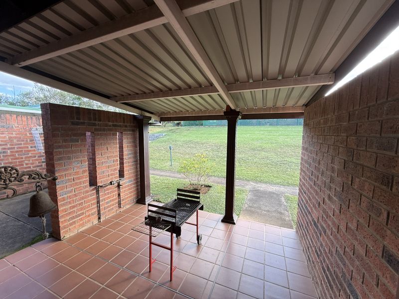 A covered outdoor patio featuring brick walls, tiled flooring, and a corrugated metal roof, overlooking a spacious rear garden area.