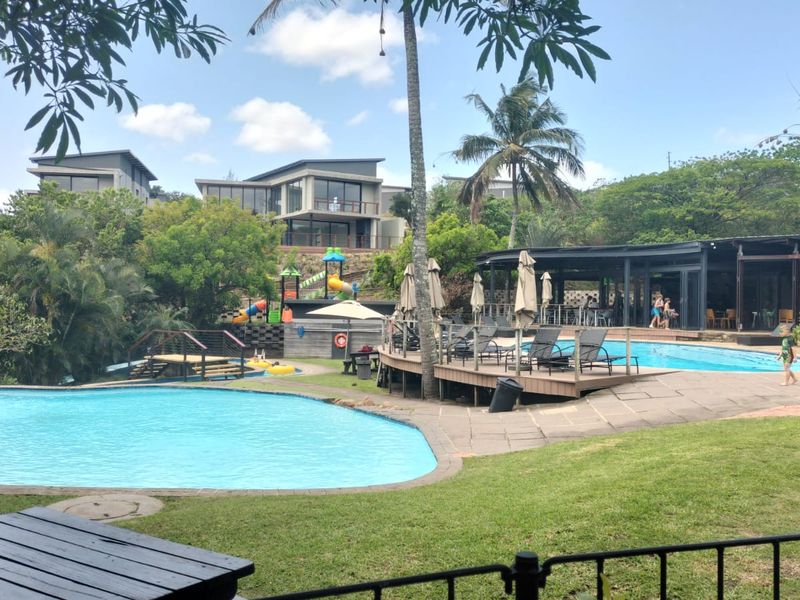 A well-maintained communal pool area featuring a swimming pool, wooden decking, paved pathways, and landscaped garden surroundings within the cluster estate.
