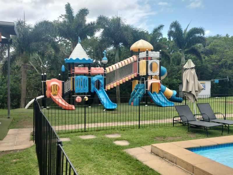 Communal pool area featuring a swimming pool, paved patio, and a children's playground set within a landscaped garden.
