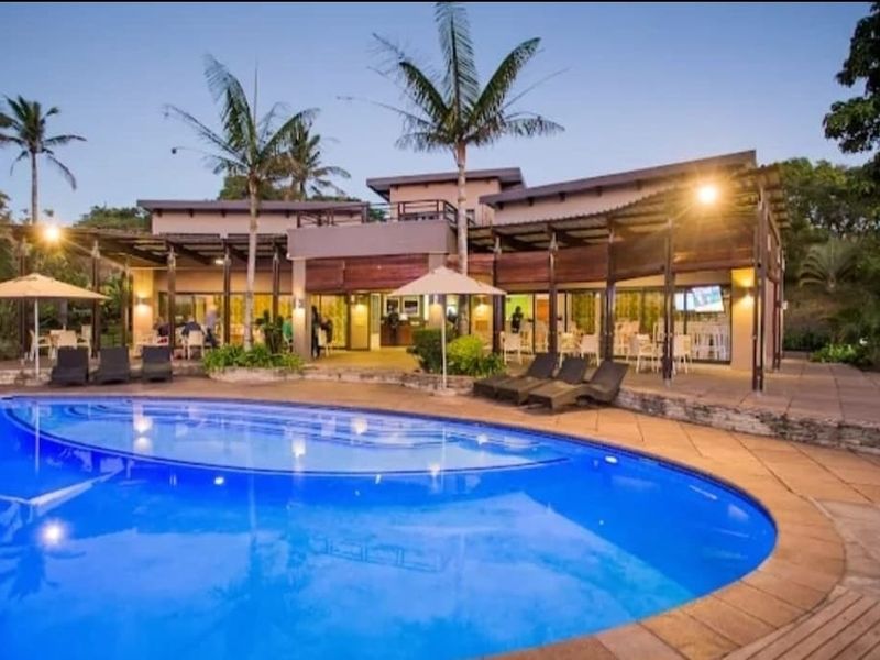 A well-maintained outdoor pool area featuring a large swimming pool, paved deck, and a covered patio area with integrated lighting.