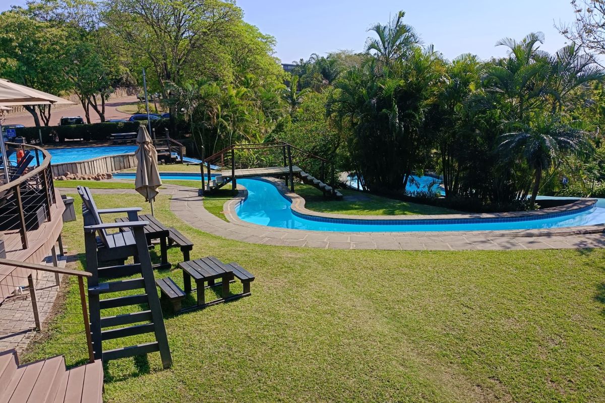 A well-maintained communal pool area featuring a winding swimming pool, wooden footbridge, landscaped garden, and timber decking for outdoor relaxation.