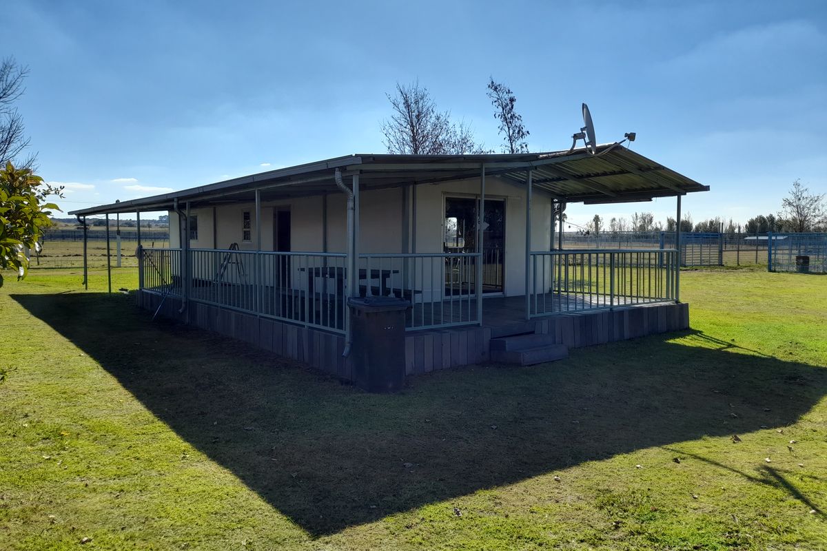 A detached cottage featuring a wrap-around veranda with metal railings, timber decking, and a corrugated metal roof set in a spacious garden.