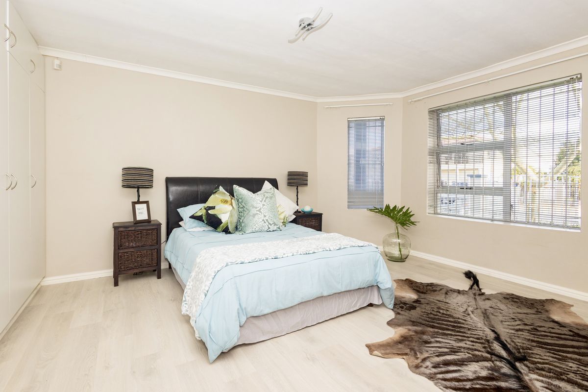 A well-lit bedroom featuring light-toned laminate flooring, built-in cupboards, and large windows with venetian blinds.