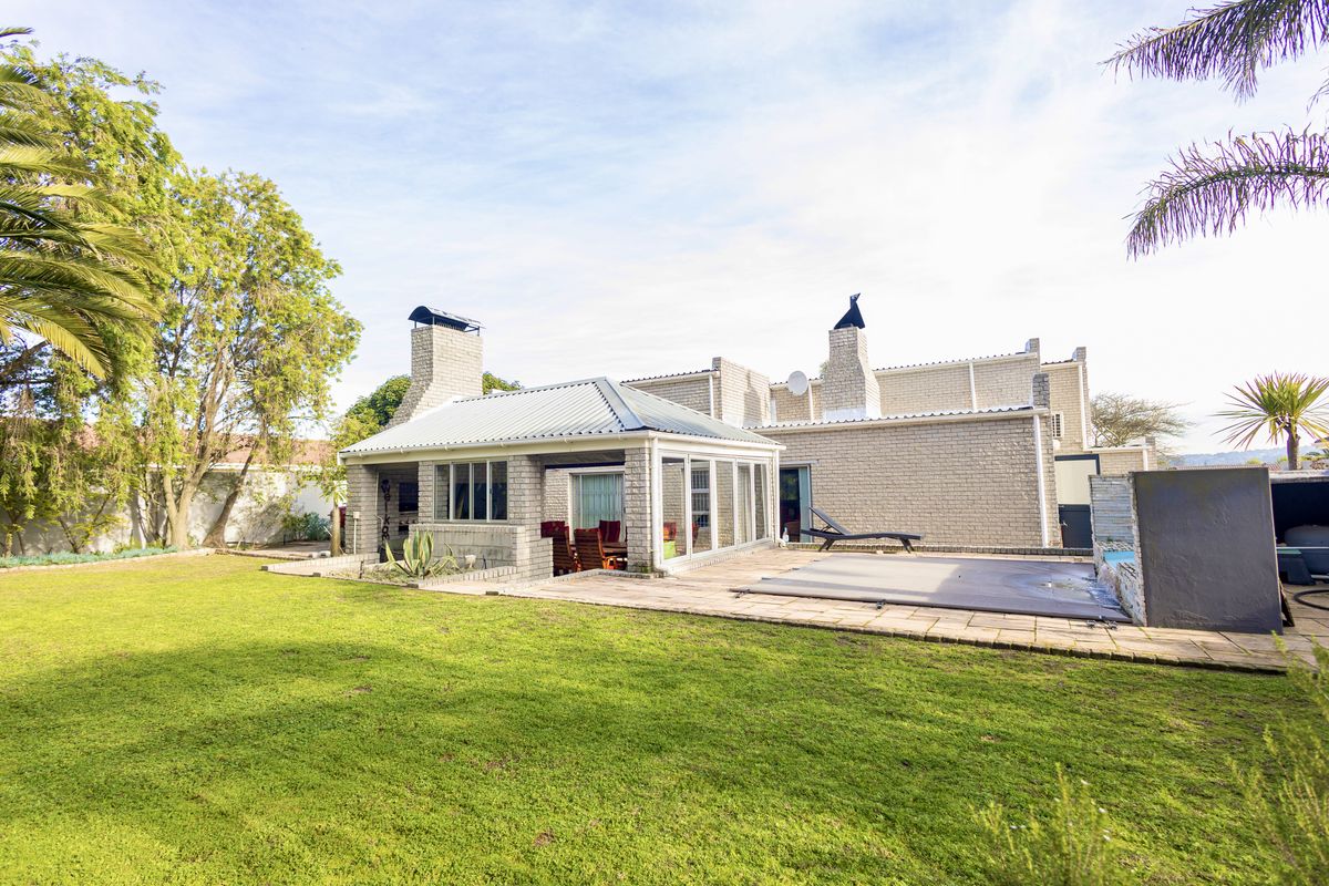 Rear view of the property featuring a spacious lawn, a covered patio with a built-in braai, and a swimming pool with a deck.