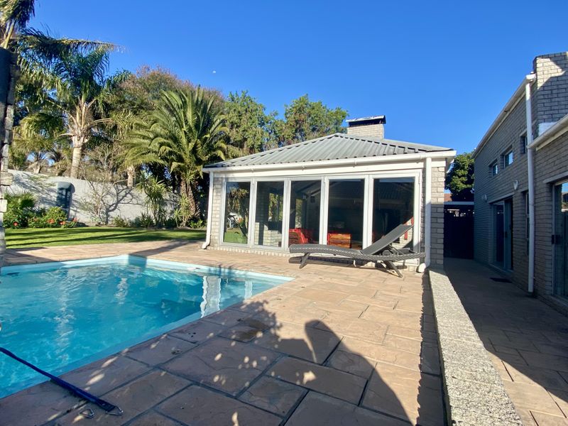 A well-maintained outdoor pool area featuring a paved deck, a covered patio with bi-fold glass doors, and a brick-clad structure.