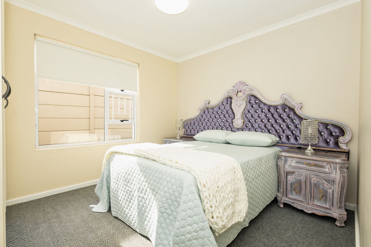 A well-maintained bedroom featuring neutral wall tones, carpeted flooring, and a functional sliding window with a roller blind.
