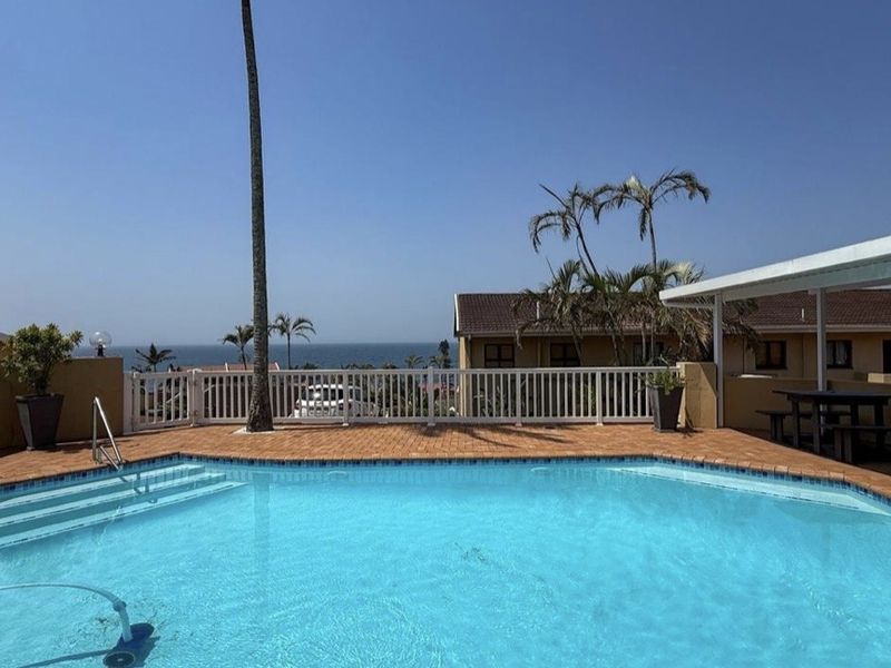 A well-maintained outdoor swimming pool area featuring brick paving, a white balustrade, and a covered patio with scenic ocean views.