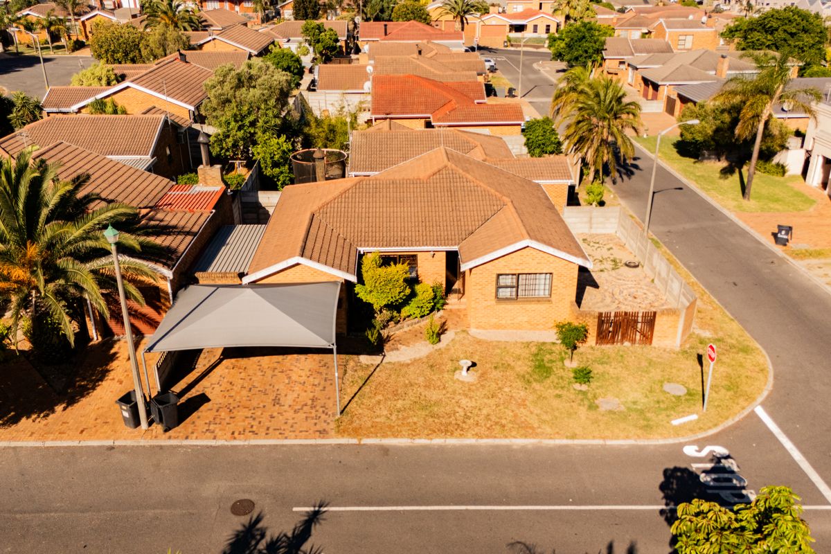 Aerial view of a residential property featuring a brick facade, tiled roof, paved driveway with a shade sail carport, and a corner lot garden.