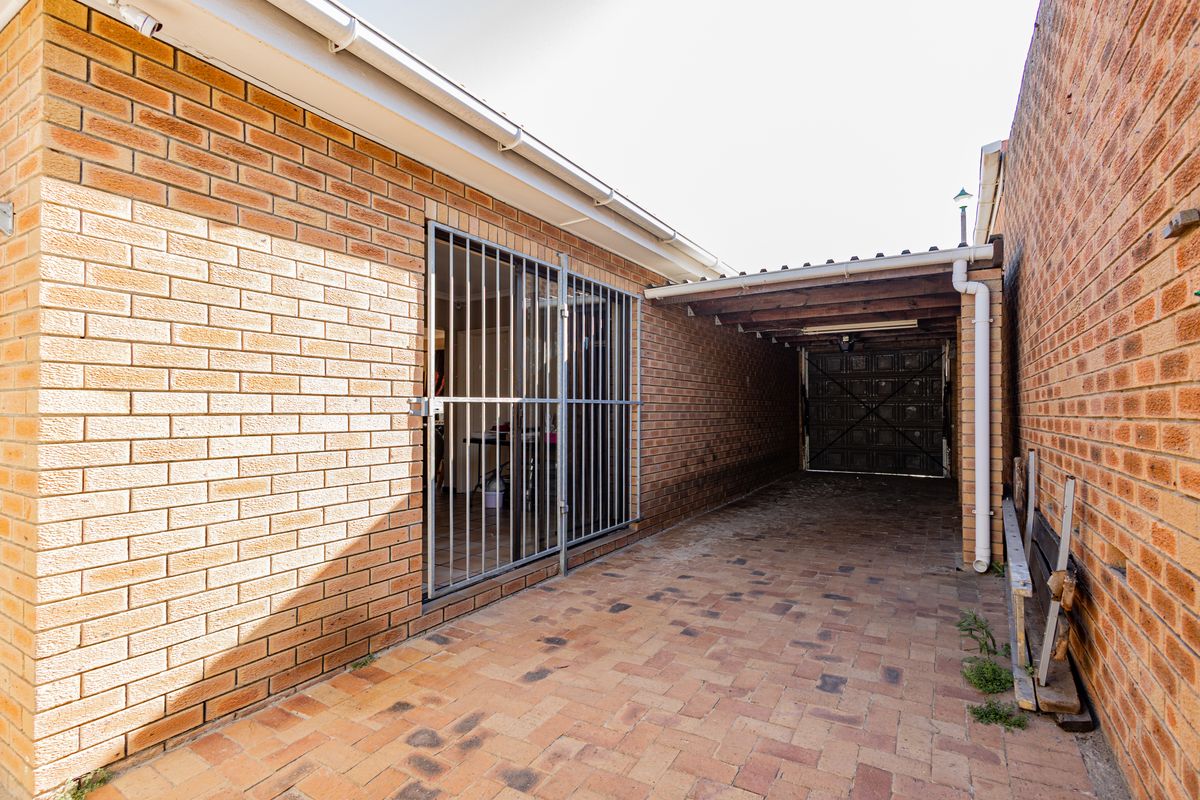 A paved rear exterior area featuring a covered carport, brick wall construction, and a secure metal gate providing access to the property.