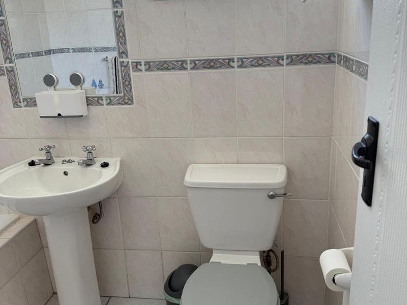 A functional bathroom featuring a pedestal washbasin, toilet, and neutral ceramic wall and floor tiling with decorative border accents.
