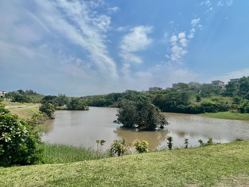 A scenic view of a tranquil lake surrounded by lush greenery and distant residential properties, showcasing the natural landscape of the estate.