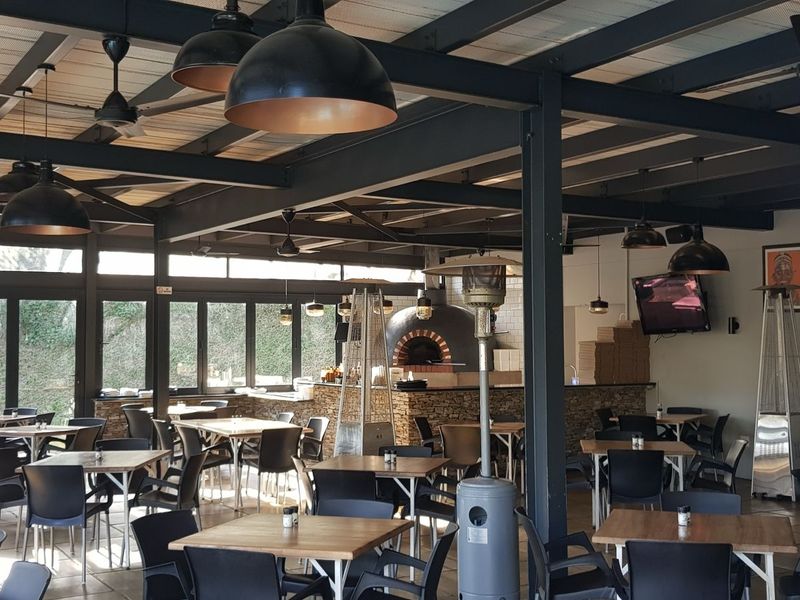 A spacious covered patio area featuring exposed steel beams, a built-in stone pizza oven, and a bar counter, ideal for outdoor dining and entertainment.