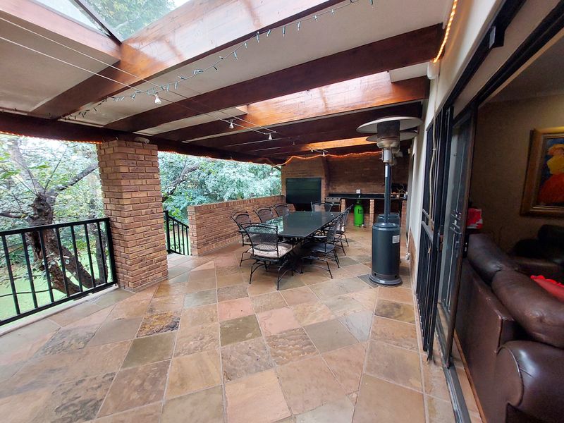 Covered outdoor patio featuring exposed timber beams, brick pillars, stone tile flooring, and a built-in braai area, perfect for alfresco dining.