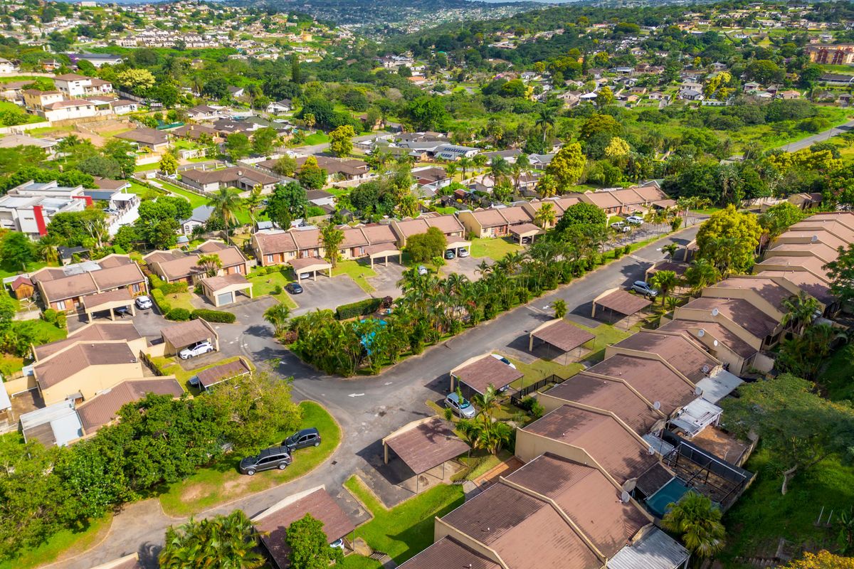An aerial view of a residential townhouse complex featuring tiled roof units, private carports, and a communal swimming pool set within a landscaped estate.