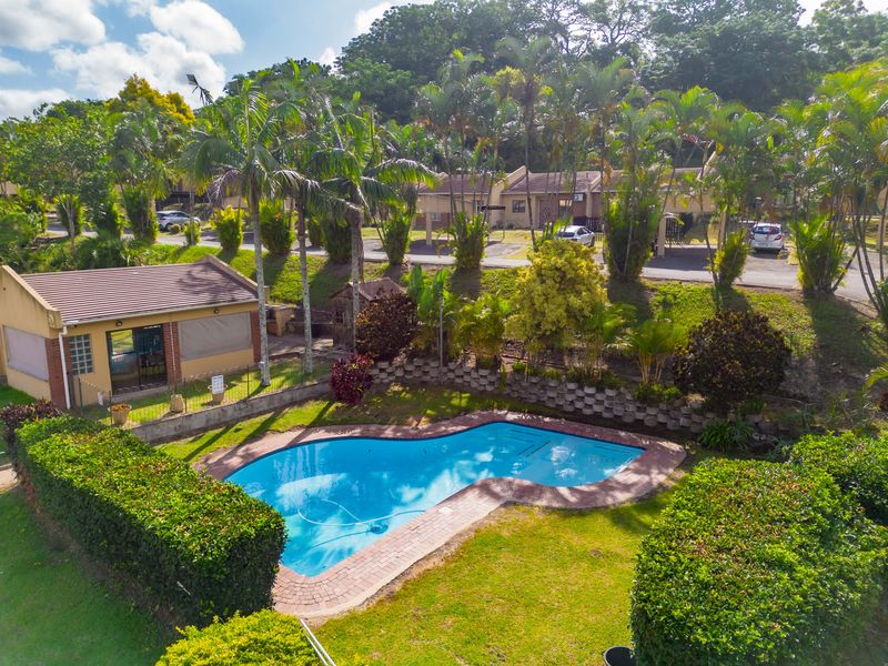 Aerial view of a private swimming pool set within a landscaped garden, featuring brick coping and a nearby outbuilding in a residential complex.