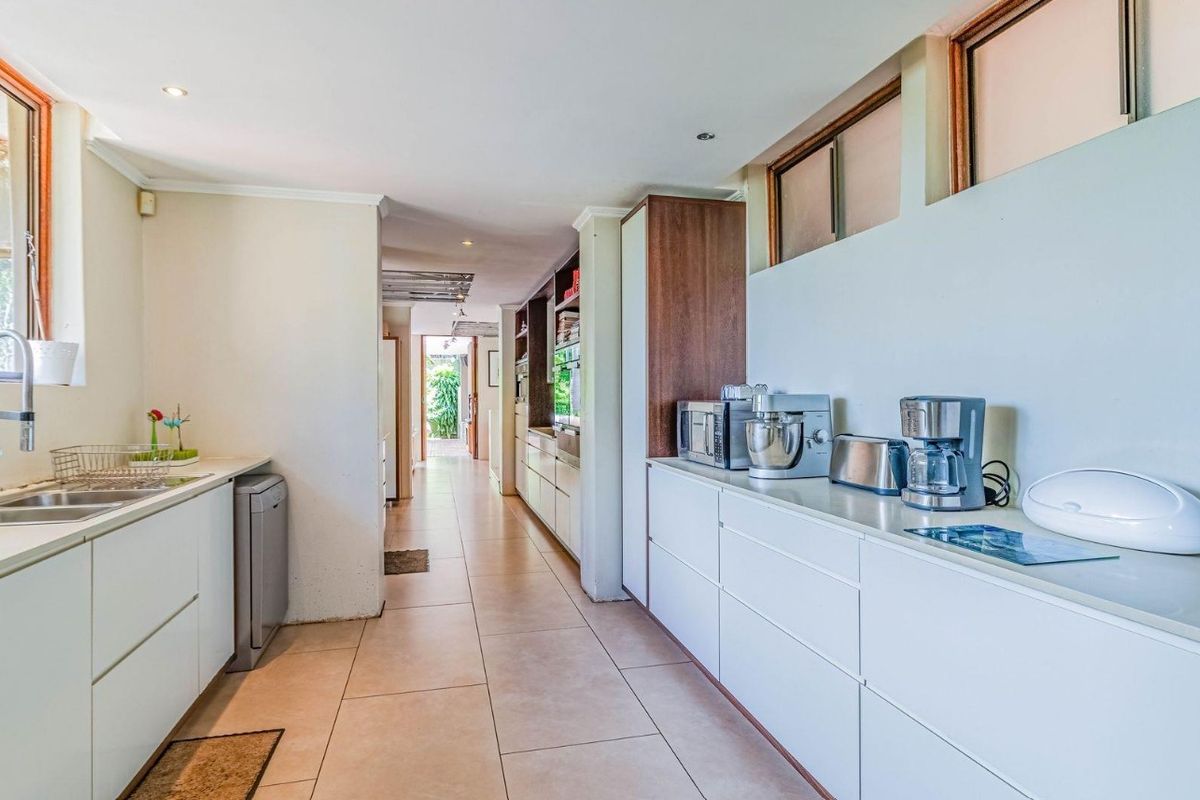 A modern galley-style kitchen featuring sleek white cabinetry, tiled flooring, double sink, and high-level windows providing natural light.