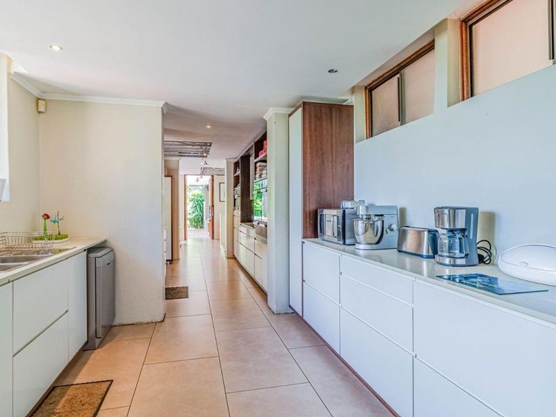 A modern galley-style kitchen featuring sleek white cabinetry, tiled flooring, double sink, and high-level windows providing natural light.