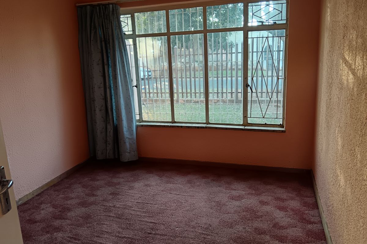 An empty bedroom featuring a large multi-pane window with security bars, textured walls, and carpeted flooring, offering a blank canvas for renovation.