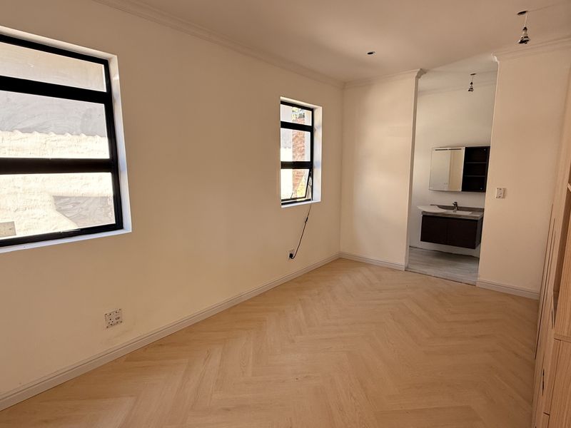 A bright, vacant bedroom featuring modern herringbone flooring, black-framed windows, and an adjoining en-suite bathroom with a contemporary vanity.