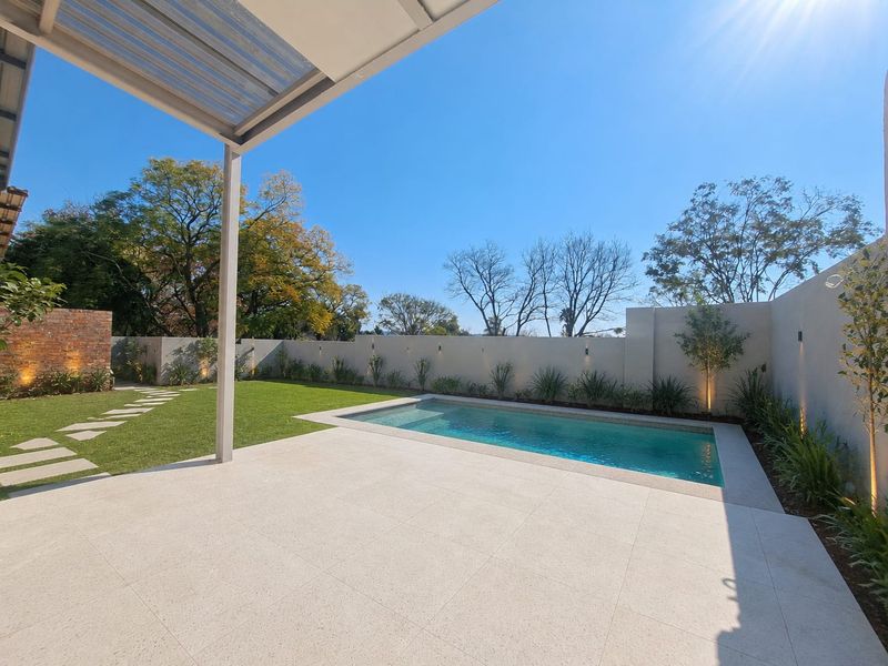 A well-maintained outdoor area featuring a swimming pool, tiled patio, covered veranda, and landscaped garden with perimeter wall lighting.