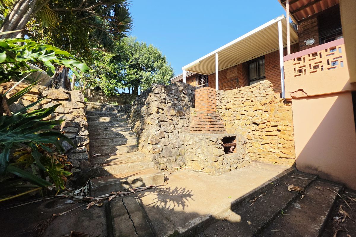 Rear exterior area featuring a stone retaining wall, built-in brick braai, concrete stairs, and a covered patio space.
