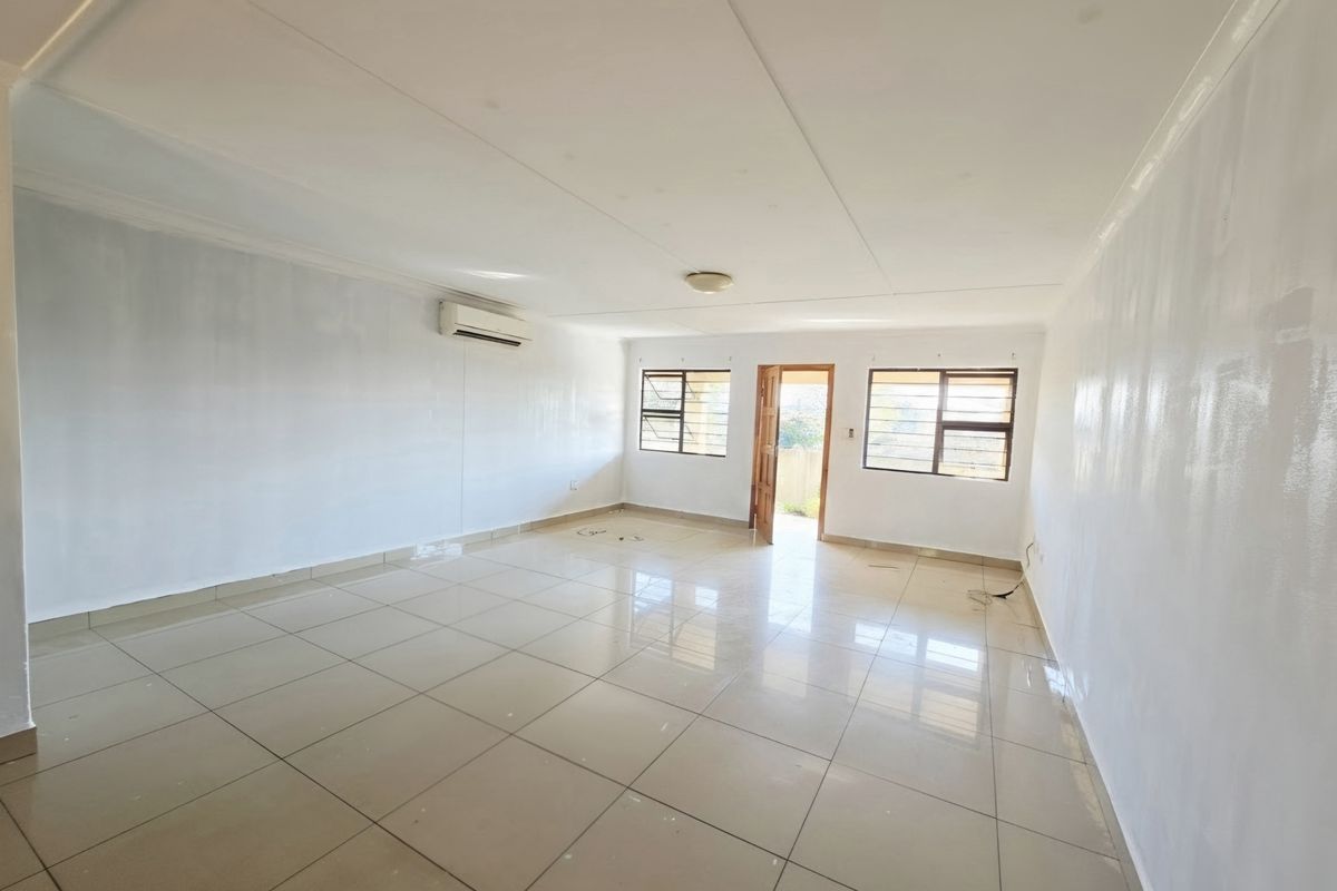 A spacious, empty living area featuring neutral tiled flooring, a wall-mounted air conditioning unit, and large windows providing natural light.