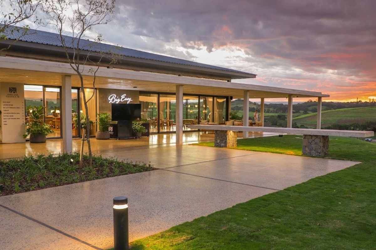 A modern clubhouse exterior featuring a spacious covered patio, expansive sliding glass doors, and high-quality finishes overlooking a scenic landscape at sunset.
