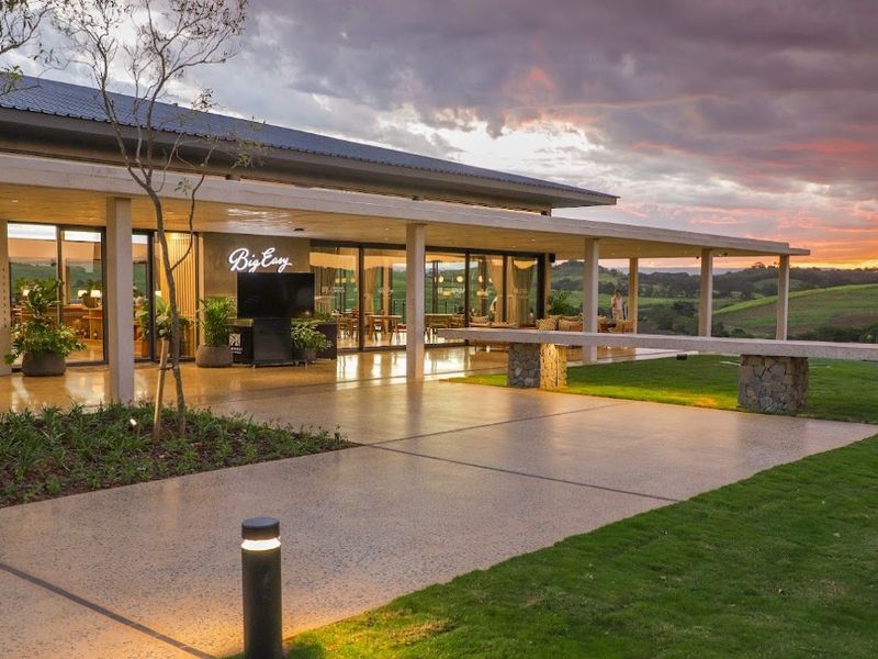 A modern clubhouse exterior featuring a spacious covered patio, expansive sliding glass doors, and high-quality finishes overlooking a scenic landscape at sunset.