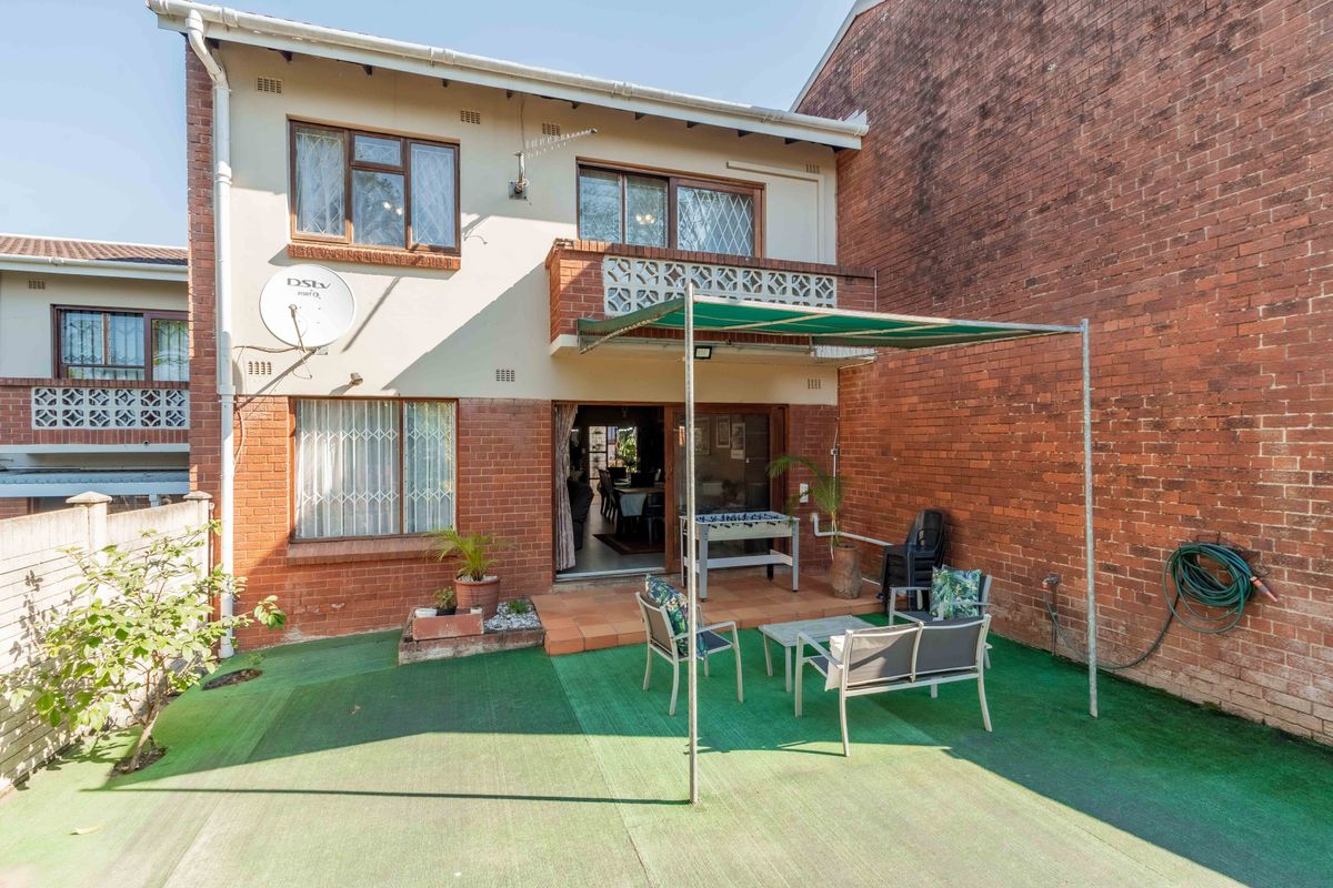 Rear view of the townhouse featuring a brick facade, a small balcony, sliding door access, and a paved patio area with a metal awning.