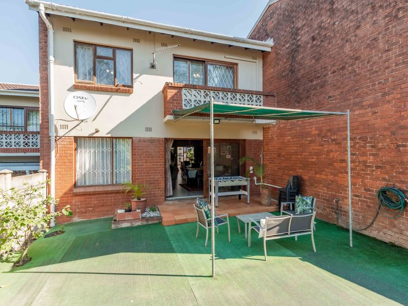 Rear view of the townhouse featuring a brick facade, a small balcony, sliding door access, and a paved patio area with a metal awning.