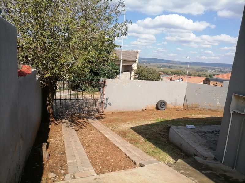 Front exterior view featuring a paved driveway, metal gate, and boundary wall with surrounding garden space and distant landscape views.