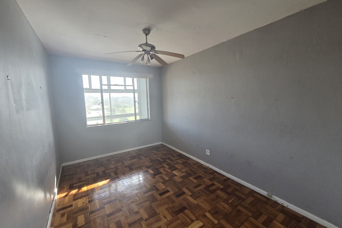 An empty bedroom featuring original parquet flooring, a ceiling fan, and large windows providing natural light. The room requires minor cosmetic updates to walls and finishes.