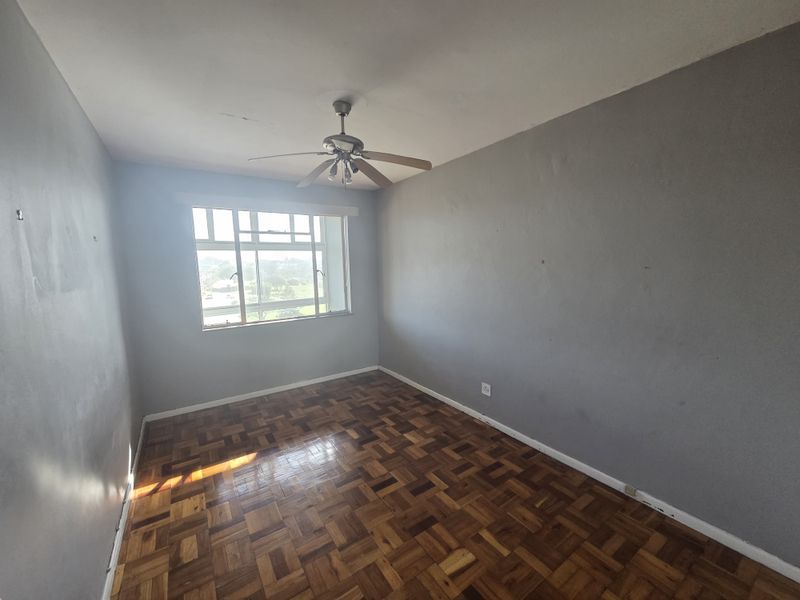 An empty bedroom featuring original parquet flooring, a ceiling fan, and large windows providing natural light. The room requires minor cosmetic updates to walls and finishes.