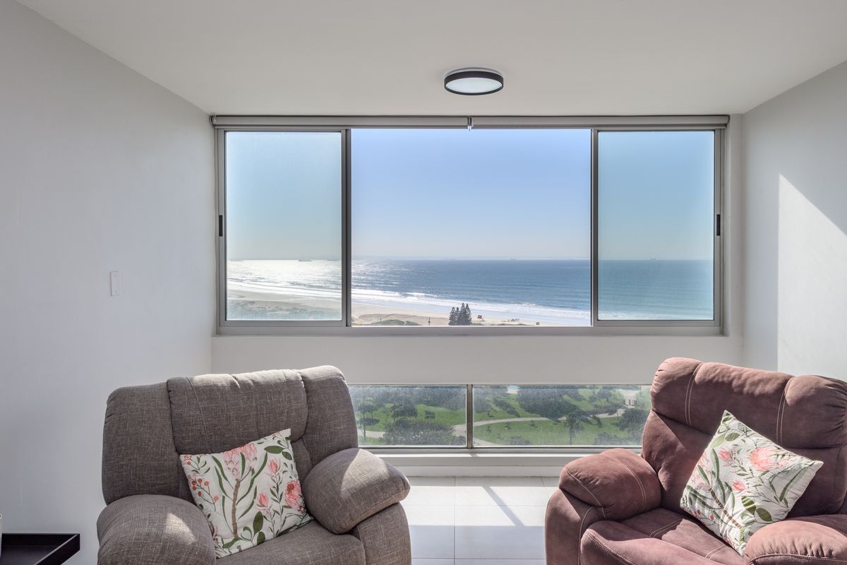 A bright living area featuring large sliding windows that frame a panoramic ocean and beach view, complemented by a modern glass balustrade.