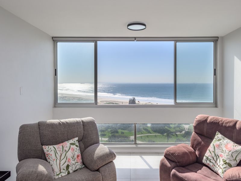 A bright living area featuring large sliding windows that frame a panoramic ocean and beach view, complemented by a modern glass balustrade.