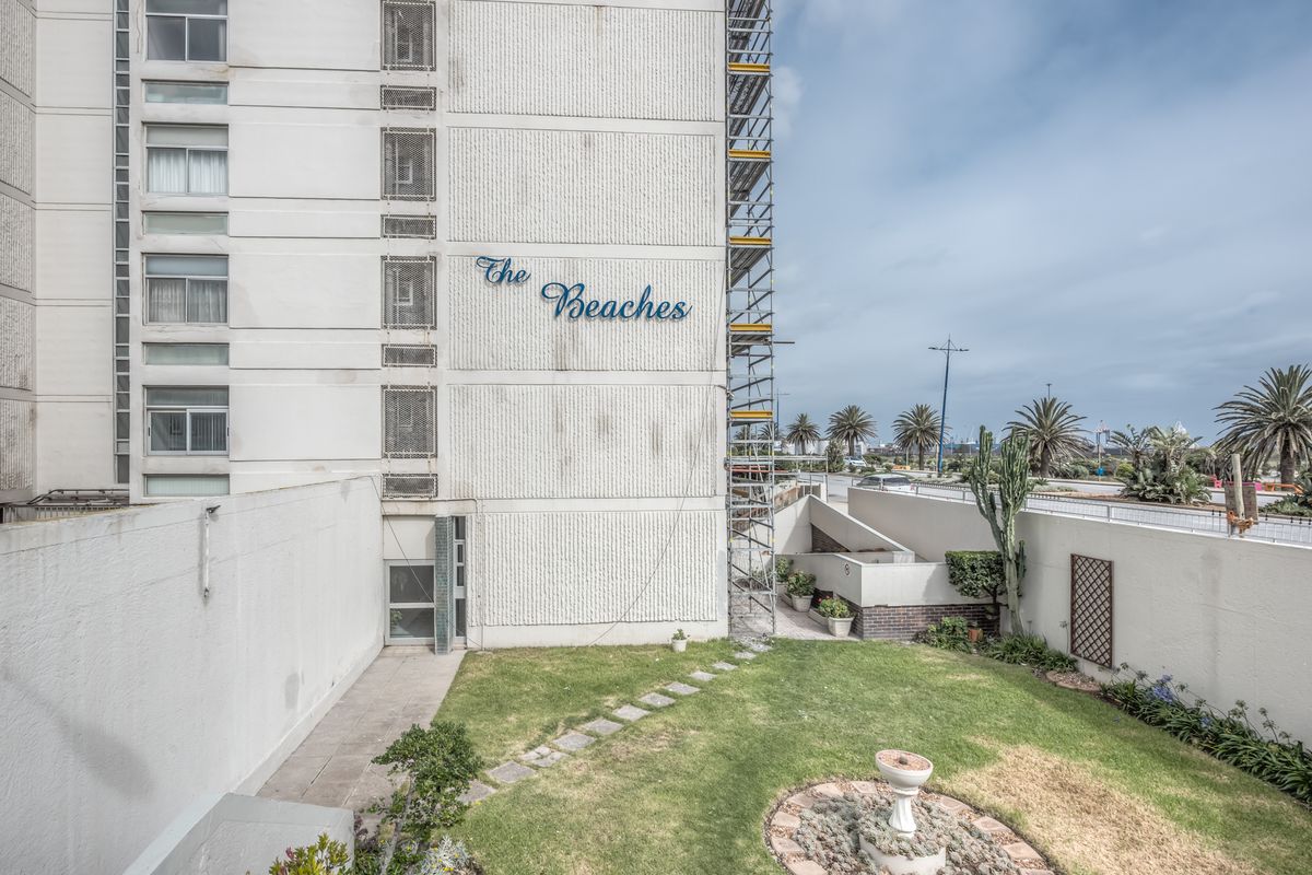 Rear exterior view of The Beaches apartment complex featuring a lawn area, stone pathway, and visible scaffolding on the building facade.