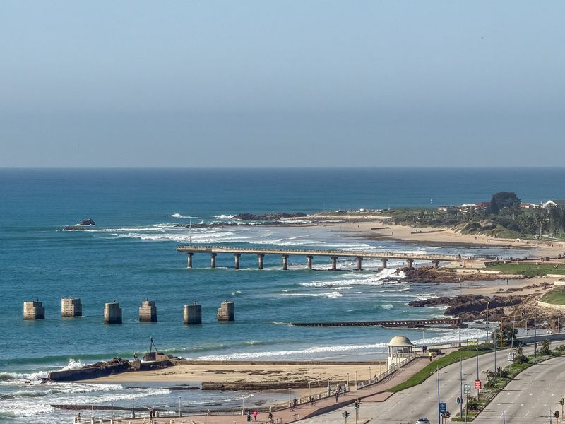 A scenic elevated view overlooking the coastline, featuring the ocean, sandy beach, and a prominent pier.