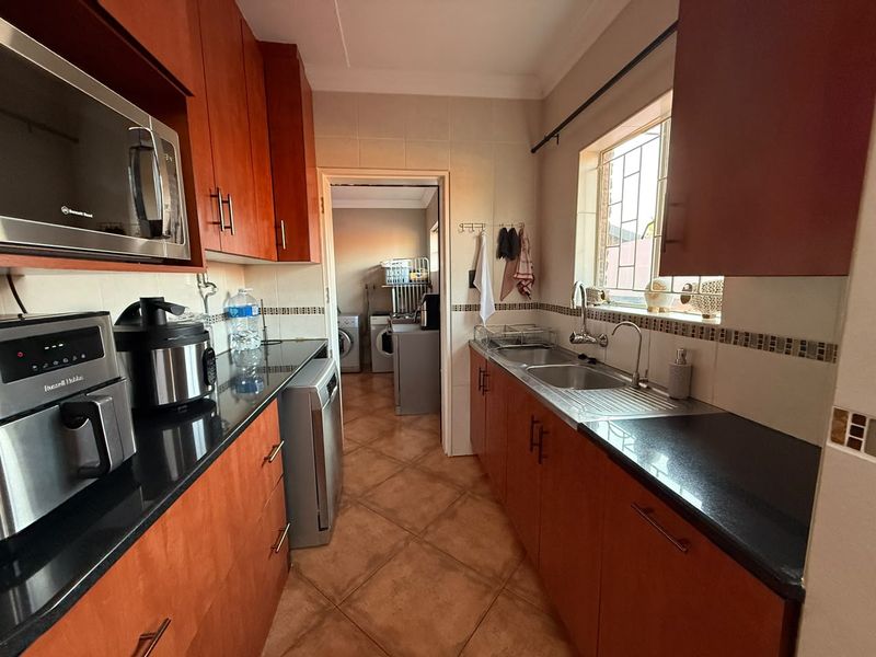 A functional galley-style kitchen featuring warm-toned wooden cabinetry, dark granite countertops, and a double basin sink with a view into the adjacent laundry area.