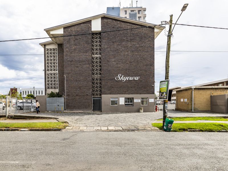 Street-facing view of the Skyview apartment building, featuring a brick facade, decorative breeze block detailing, and a paved entrance area with security fencing.