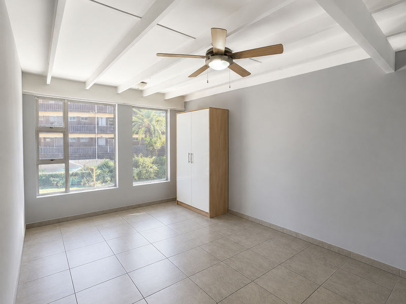 A bright, spacious bedroom featuring exposed ceiling beams, a ceiling fan, neutral tiled flooring, and a built-in wardrobe.