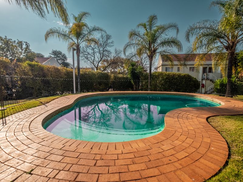 A well-maintained kidney-shaped swimming pool featuring brick coping, surrounded by a manicured garden and secure metal fencing.
