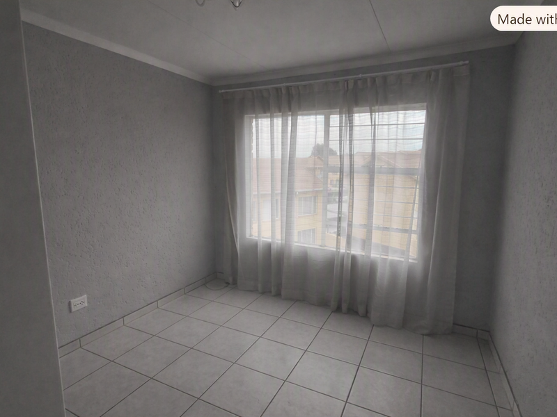 A vacant bedroom featuring neutral tiled flooring, textured walls, and a window fitted with security bars and sheer curtains.