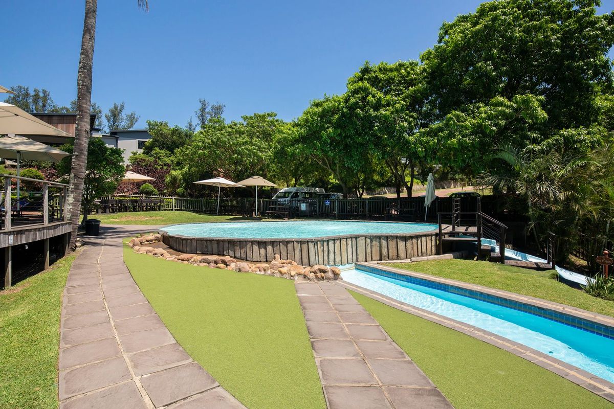 A well-maintained outdoor pool area featuring a circular swimming pool, a water slide, paved walkways, and lush surrounding garden landscaping.