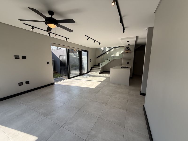 Modern open-plan living area featuring large format floor tiles, track lighting, a sleek staircase with glass balustrade, and a central kitchen island.