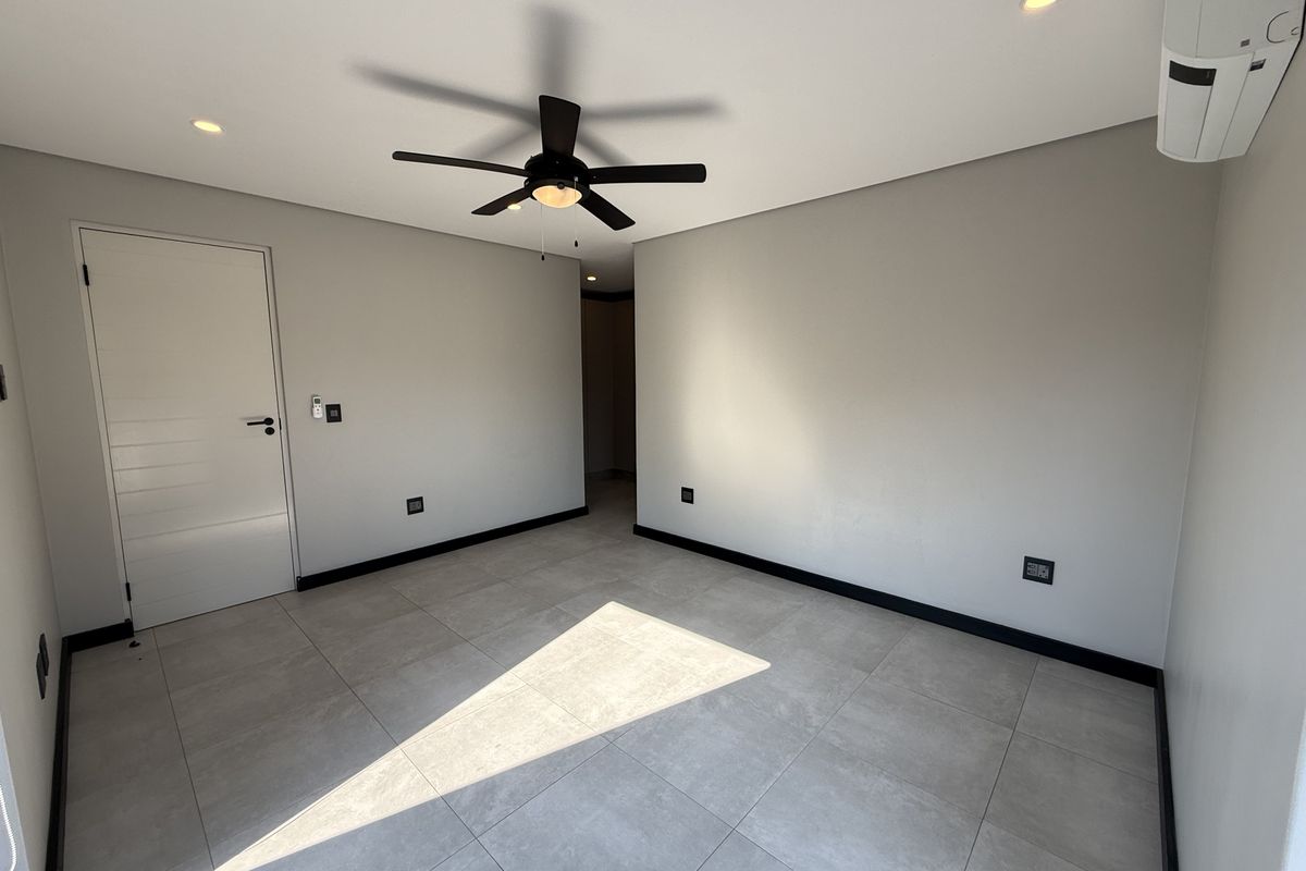 A bright, modern, and empty bedroom featuring neutral tiled flooring, recessed lighting, a ceiling fan, and a wall-mounted air conditioning unit.